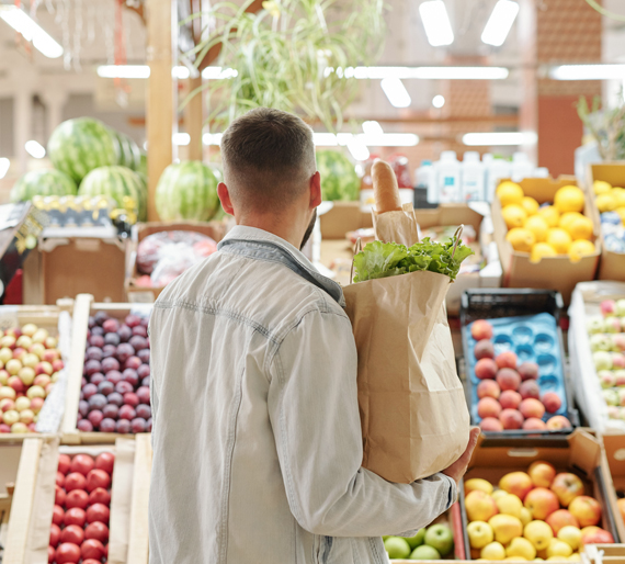 Hombre de espaldas haciendo la compra en un mercado con una bolsa de papel llena de alimentos.